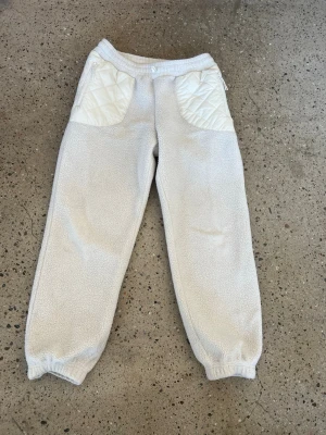 Beiga fleecebyxor med quiltade detaljer - Mysiga beiga mjukisbyxor i teddyfleece med quiltade fickor framtill och dragkedja. Resår i midja och benslut för skön passform. Perfekta för chill eller streetwear-stil. Materialet är mjukt och värmande, och detaljerna ger en unik look. Oversized!!