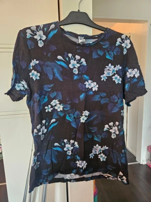 Svart blommig t-shirt med blå toner - Snygg svart t-shirt med korta ärmar och blommigt mönster i vitt och blått. T-shirten har en avslappnad passform och rund halsringning. Perfekt för dig som gillar stilrena men ändå färgglada plagg.