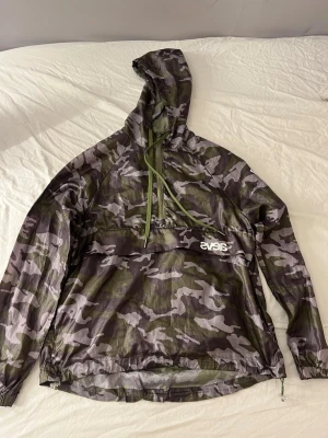 Camouflage vindjacka från Svea - Snygg vindjacka från Svea i camouflage-mönster med dragkedja och huva. Jackan har en stor ficka framtill med Svea-logga och justerbar nederkant. Tillverkad i lätt och tunt material, perfekt för blåsiga dagar.