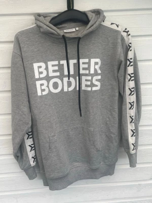 Grå Better Bodies hoodie med tryck - Snygg grå hoodie från Better Bodies med stor vit logga på bröstet och svarta snören i huvan. Ärmarna har vita band med svarta mönster längs sidorna. Klassisk känguruficka framtill och ribbade muddar. Perfekt för en sportig och avslappnad stil.