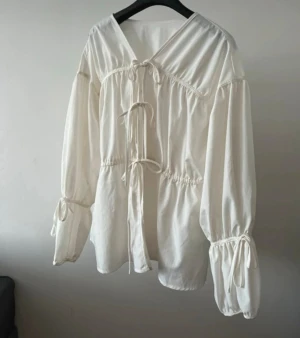 Vit blus med knytband och v-ringning - Stilren vit blus med v-ringning och dekorativa knytband framtill. Blusen har långa ärmar med resår och knyt vid handlederna för en trendig look. Tillverkad i ett lätt och luftigt tyg som ger en avslappnad känsla.