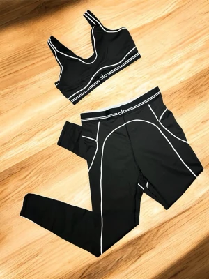 Svart set från Alo med vita detaljer - Snyggt svart underklädesset från Alo med sport-bh och matchande tights. Setet har vita kontrastsömmar och logga på både bh och midja. Materialet är stretchigt och följsamt, perfekt för en aktiv livsstil eller som bekväma loungekläder.