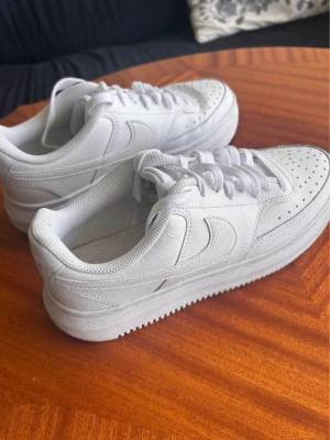 Vita Nike Air Force 1 sneakers - Fräscha vita Nike Air Force 1 sneakers med klassisk siluett, perforerad tå och tydlig swoosh på sidorna. Skorna har lågt skaft, tjock sula och är tillverkade i läder och textil. Perfekta för en clean och stilren look.