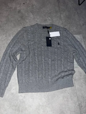 Grå kabelstickad tröja Polo Ralph Lauren - Snygg grå kabelstickad tröja från Polo Ralph Lauren med rund halsringning. Den är oanvänd och ny samt får du original förpackningen med. 