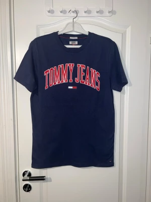 Blå Tommy Jeans t-shirt - Snygg marinblå t-shirt från Tommy Jeans med stort rött och vitt tryck på bröstet. Klassisk rund halsringning och korta ärmar. Perfekt för en avslappnad och trendig look. Finns att hämta i Södertälje.