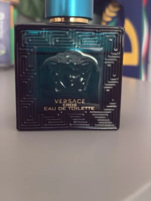 Versace Eros Eau de Toilette - Versace Eros Eau de Toilette i en elegant turkos glasflaska med Medusa-huvudet i relief och guldtext. Flaskan har en fyrkantig form med grekiskt mönster och blått metallock. En ikonisk doft för dig som gillar exklusiva parfymer.