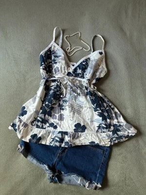 Blommig sommartopp från Hilfiger Denim - Blommig topp från Hilfiger Denim i vitt och marinblått. Toppen har smala axelband, v-ringning, knyt i midjan och volangkant nedtill. Spetts längst ringningen och perfekt för sommaren i 100% bomull. Använd och är missfärgad på insidan av axelbanden (se bild) men syns inte alls när man har den på sig ☀️