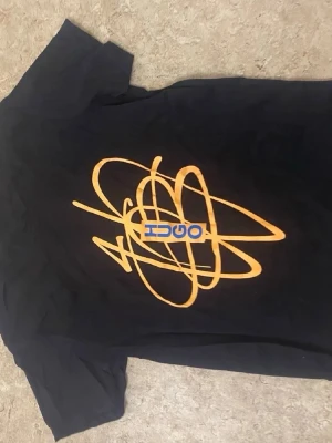Svart t-shirt med orange tryck OSCI - Cool svart t-shirt från OSCI med ett stort orange grafiskt tryck på ryggen och blå textdetalj. Klassisk passform och korta ärmar, perfekt till jeans eller shorts. Tillverkad i mjukt material för skön känsla hela dagen.