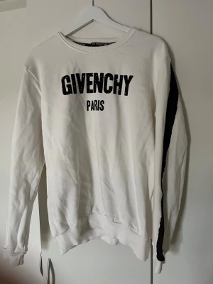 Vit sweatshirt från Givenchy Paris - L men sitter som M!! Säljer en vit sweatshirt från Givenchy Paris med svart logotyptryck framtill och svarta ränder längs ärmarna. Tröjan har rund halsringning, ribbade muddar och är tillverkad i mjukt material. Perfekt för en trendig och avslappnad stil.