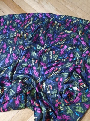 Mörkblå scarf med färgglada flygplan - Snygg och unik scarf i mörkblått med färgglada flygplansmotiv i neonfärger som rosa, gul, grön och blå. Tunt och mjukt material som ger en cool vibe till din outfit. Perfekt accessoar för dig som gillar statement-plagg och lekfulla mönster.