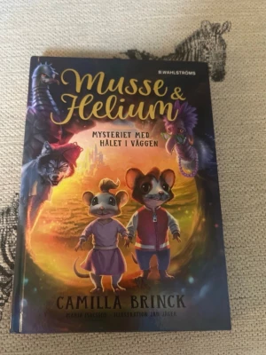 Musse & Helium: Mysteriet med hålet i väggen - En färgglad och fantasifull bok av Camilla Brinck om mössen Musse och Helium som dras in i ett magiskt äventyr genom ett hål i väggen. Boken har ett hårt omslag med illustrationer av djur och magiska miljöer. Perfekt för dig som gillar spännande och roliga berättelser.