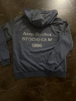 Blå hoodie från Acne Studios M - Snygg blå hoodie från Acne Studios med tryck på ryggen: 'Acne Studios STOCKHOLM 1996'. Klassisk modell med huva och ribbade muddar. Perfekt för en avslappnad och trendig stil. Materialet känns mjukt och skönt mot huden.