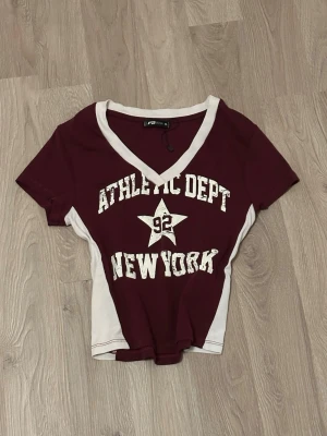 En vin röd topp från new yorker - Säljer en vinröd croppad topp från F&F med vit v-ringad kant och texten 'ATHLETIC DEPT 92 NEW YORK' i vitt framtill. Toppen har korta ärmar och är gjord i ett stretchigt bomullsmaterial. Perfekt för en sportig och trendig look.