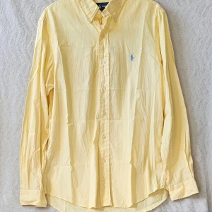 Gul skjorta från Ralph Lauren - Klassisk gul skjorta från Ralph Lauren med blå broderad logga på bröstet. Skjortan har lång ärm, knappar framtill och en avslappnad passform. Tillverkad i mjuk bomull och perfekt för en stilren look.