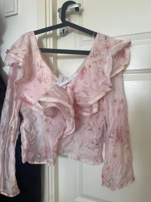 Rosa blommig volangblus Bubbleroom - Supersöt blus från Bubbleroom i tunn, transparent rosa mesh med blommigt mönster. Blusen har stora volanger längs ringningen och långa ärmar. Storlek S. Prislapp kvar!