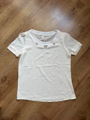 Vit blus med cut out-detaljer - Snygg vit blus med korta ärmar och unika cut out-detaljer vid halsringningen. Blusen har en rak passform och är tillverkad i ett lätt, tunt material som känns luftigt. Perfekt för dig som gillar stilrena och moderna toppar.