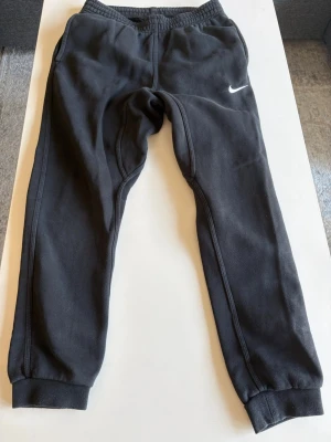 Svarta mjukisbyxor från Nike stl S - Säljer ett par svarta mjukisbyxor från Nike med klassisk vit swoosh-logga på vänster ben. Byxorna har resår i midjan och vid bensluten samt sidofickor. Tillverkade i mjukt och bekvämt material, perfekta för chill dagar.