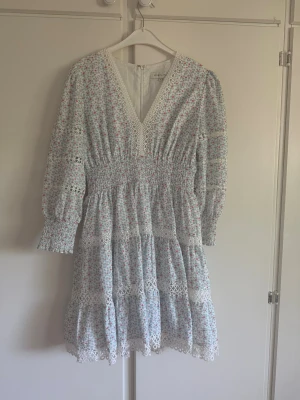 Blommig klänning från By Malina - Supersöt blommig klänning från By Malina med v-ringning, spetsdetaljer och trekvartsärm. Klänningen har smock i midjan och volangkant nertill. Färgerna går i vitt, blått och rött med små blommor. Perfekt för sommarens alla tillfällen!