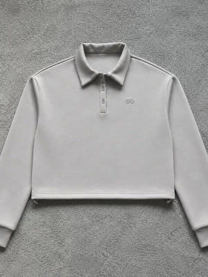 Grå cropped sweatshirt från Alo - Stilren grå cropped sweatshirt från Alo med klassisk krage och knappslå framtill. Tröjan har långa ärmar, ribbade muddar och minimalistisk logga broderad på bröstet. Perfekt för en avslappnad och trendig look.