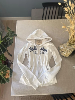 Vit hoodie från Hollister med dragkedja - Säljer en vit hoodie från Hollister med dragkedja framtill.