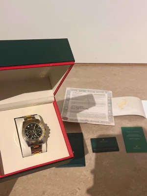 Role Daytona guld och silver herrklocka - Säljer en lyxig Role Daytona herrklocka med svart urtavla och tre kronografer. Armbandet är i tvåtonad design med både guld och silver. Klockan levereras i originalbox med certifikat och tillbehör. Perfekt för dig som vill sticka ut med stil.