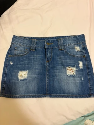 Blå jeanskjol med slitningar - Snygg blå jeanskjol i kort modell med slitna detaljer framtill och bakfickor. Kjolen har bälteshällor, knappstängning och en liten hjärtformad detalj bak. Perfekt för en trendig och avslappnad look.