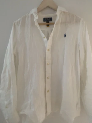 Vit linneskjorta från Polo Ralph Lauren - Klassisk vit skjorta i linne från Polo Ralph Lauren med lång ärm och knappar framtill. Skjortan har den ikoniska broderade loggan på bröstet och en avslappnad passform. Perfekt till sommaren för en clean och fräsch look.☀️⭐️