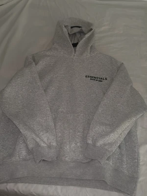Grå Essentials Fear of God hoodie - Snygg grå hoodie från Essentials Fear of God med svart logga tryckt på bröstet och ryggen. Klassisk passform med huva och ribbade muddar. Perfekt för en avslappnad och trendig streetwear-look.