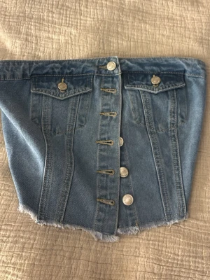 Kort jeanskjol med knappar - Säljer en kort jean tubbtopp i ljusblå denim med fransig kant nertill och dekorativa fickor framtill. Kjolen har knappar längs hela framsidan och klassiska sömmar för en cool look.