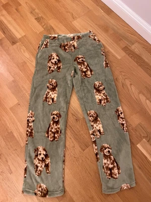 pyjamasbyxor  - Mysiga pyjamasbyxor i mjuk fleece med gröngrå bas och gulligt hundmönster i brunt och beige. Byxorna har rak passform och resår i midjan för extra komfort. Perfekta för slappa dagar hemma eller när du vill ha det extra bekvämt. Köpt på Gekås Ullared 