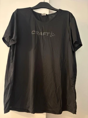 Svart träningstopp från Craft - Säljer en svart träningstopp från Craft med rund halsringning och korta ärmar. Tillverkad i ett lätt och ventilerande funktionsmaterial som passar perfekt till träning. Diskret logga på bröstet i silver. Enkel och sportig design. Storlek XXL.