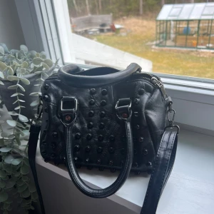 Svart handväska med nitar - Snygg svart handväska i skinn med coola nitar på framsidan. Väskan har både handtag och en avtagbar axelrem, man får plats med mycket i den💕Har ett litet slitage som inte syns då det är lagat. Skriv för bild på det om ni undrar