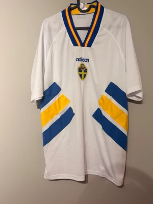 Svensk landslagströja Adidas vit - Vit svensk landslagströja från Adidas med blå och gula detaljer samt broderat landslagsmärke på bröstet. Tröjan har en V-ringad krage med blå och gula ränder och korta ärmar. Perfekt för fotbollsfans som vill visa sitt stöd.