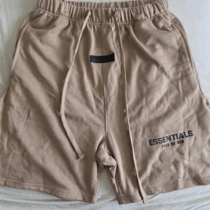 Beige shorts från Essentials Fear of God - Säljer ett par beige shorts från Essentials Fear of God med långa snören i midjan och tryckt logga på vänster ben. De har en avslappnad passform och elastisk midja för extra komfort. Perfekta för sommardagar eller chill hemma.