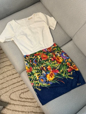 Vit och blommig klänning från Siva - Superfin klänning från Siva exclusive med vit överdel och kortärmad design. Kjolen är blå med färgglatt blommönster i gult, rött, lila och grönt. Klänningen har dragkedja i sidan och är perfekt för sommarens alla tillfällen.