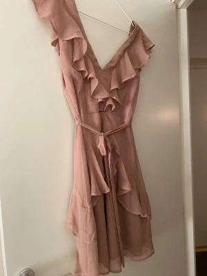 Klänning med volang  - Rosa kkänning med volangdetaljer längs v-ringningen och axlarna. Klänningen har ett knytband i midjan och är tillverkad i ett lätt, flödande material som ger en romantisk känsla. Perfekt för sommarens alla tillfällen!