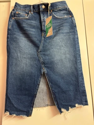 Blå jeanskjol med slits och rå kant - Trendig blå lång jeanskjol från Gina tricot, slits framtill och rå, fransig kant nedtill. Klassisk femficksmodell med knapp och dragkedja. Perfekt för dig som gillar en avslappnad men snygg denimlook.