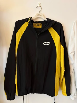 Corteiz Windbreaker - Snygg svart vindjacka med gula paneler från Corteiz. Jackan har huva, dragkedja framtill och dragsko i nederkant. Märkeslogga på bröstet och sportig design. Perfekt för dig som gillar streetwear och vill sticka ut med färg.