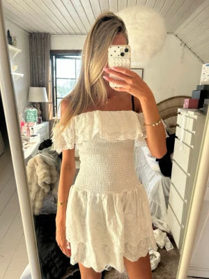 Vit offshoulder klänning med volang - Söt vit kortklänning i bomull med broderade detaljer och volang upptill. Offshoulder-modell med smockad midja och axelbandslös design. Perfekt för sommarens alla tillfällen och ger en romantisk vibe.