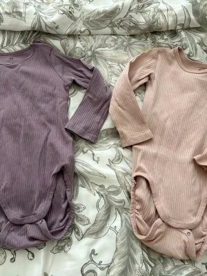 2-pack ribbade bodys från H&M - Två långärmade bodys från H&M i ribbad ekologisk bomull. En i ljuslila och en i ljusrosa, båda med tryckknappar i grenen och vid axeln för enkel påklädning. Perfekta för en bekväm och stilren vardag för de minsta.