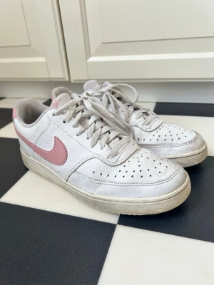 Nike Court Vision vita sneakers rosa swoosh - Nike Court Vision sneakers i vitt skinn med ljusrosa swoosh och detaljer. Klassisk låg modell med perforerad tå för ventilation och vita skosnören. Snygg retrostil som passar till det mesta och ger en clean look. Med skorna får men ett crease skydd för att skydda skon och ge den ett längre och snygg utsida Skorna är varsamt använda teken på användning se bilder 😊