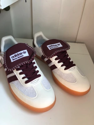 Adidas Wales Bonner sneakers vit/vinröd  - Snygga Adidas Wales Bonner sneakers i vitt och vinrött med klassiska ränder på sidan och brun gummisula. Skorna har en unik design med kontrastsömmar och bred plös med Wales Bonner-logga. Materialmix av läder och mocka för en stilren look.