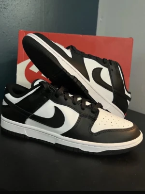 Nike Dunk Low svart/vit sneakers - Snygga Nike Dunk Low sneakers i svart och vitt läder med klassisk Swoosh-logga på sidorna. Skorna har lågt skaft, svart yttersula och vita paneler som kontrasterar mot de svarta detaljerna. Perfekta för en streetwear-look.