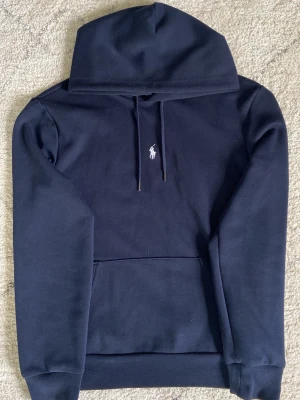 Mörkblå hoodie från Polo Ralph Lauren - Snygg mörkblå hoodie från Polo Ralph Lauren med klassisk huva och dragsnören. Ikoniska vita Polo-loggan broderad på bröstet. Hoodien har en stor magficka och ribbade muddar vid ärmar och nederkant. Tillverkad i mjukt material för maximal komfort.