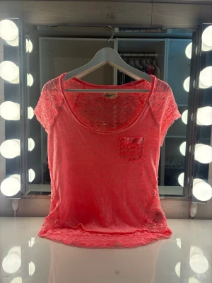 Korallrosa/neonrosa  spetstop från Hollister i strl S - Superfin korallrosa/neonrosa top med korta ärmar och rund halsringning. Toppen har spetsdetaljerpå ärmarna och hela ryggen.  Perfekt för dig som gillar en feminin och trendig look.