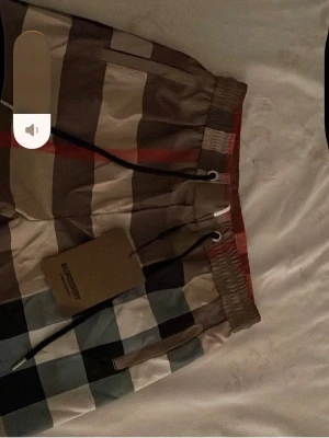  Burberry shorts - Säljer ett par klassiska rutiga shorts från Burberry med snörning i midjan. Shortsen har stora rutor i beige, brun, svart, vit och blå samt en diskret röd linje. Perfekta för sommaren och har en avslappnad passform.
