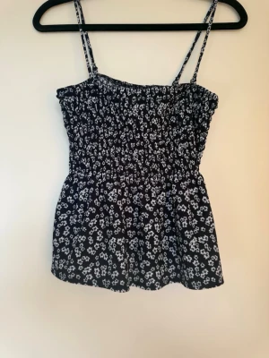Blommig svart linnetopp Gina Tricot - Supersöt svart linnetopp från Gina Tricot med vita små blommor och smock över hela toppen. Tunna axelband och lätt utsvängd passform gör den perfekt för varma dagar. Perfekt att styla med jeans eller kjol för en chill look.