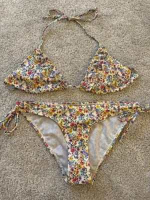 Blommig bikini  - Söt bikini med blommigt mönster i rosa, gul, blå och lila på ljus botten. Överdelen är trekantsmodell med knyt i nacken och ryggen, och underdelen har knyt i sidorna. Perfekt för sommardagar på stranden.