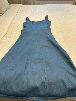 Blå jeansklänning med knytband - Snygg blå jeansklänning med axelband som knyts upptill. Klänningen har en rak och enkel design som ger en avslappnad men ändå stilren look. Perfekt för dig som gillar denim och vill ha något bekvämt och trendigt.