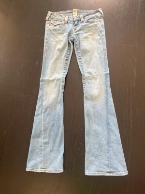 Ljusblå bootcut jeans från True Religion - Säljer ett par ljusblå bootcut jeans från True Religion med klassisk femficksdesign och sömmar. Jeansen har låg midja och utsvängda ben, perfekta för en retroinspirerad look. Tillverkade i mjuk denim med snygg tvätt o lite stretch. Inköpta i USA. 